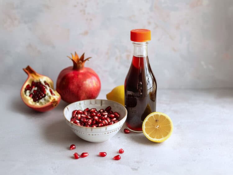 Bottle of homemade grenadine surrounded by a bowl of pomegranate seeds and half a lemon