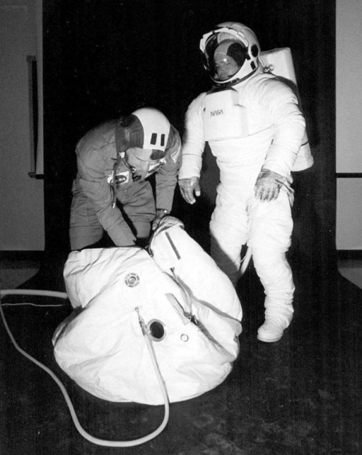 Two astronauts in space suits demonstrating how to get into the fabric rescue ball.